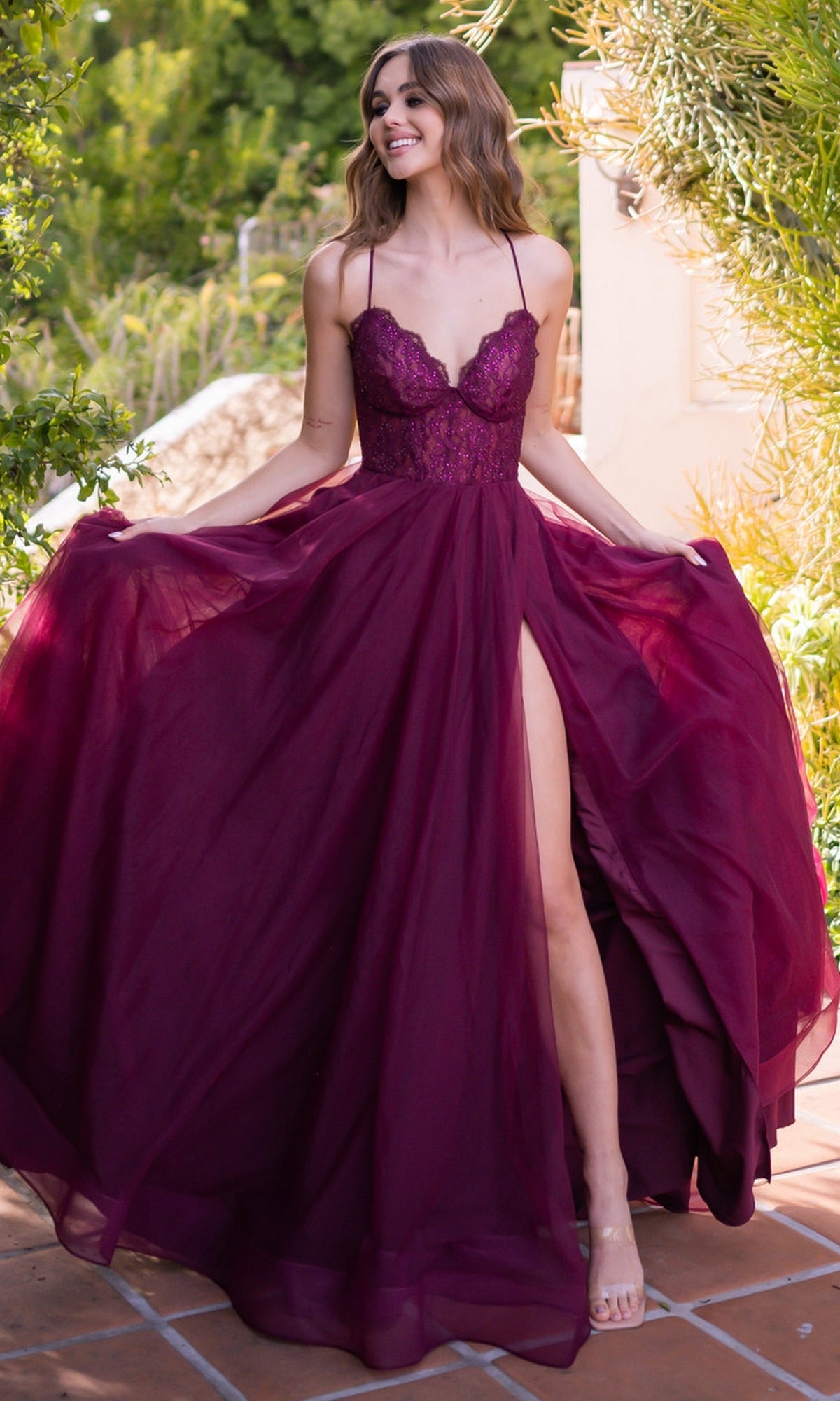 A woman in the Velvi Long Sweetheart A-Line Prom Dress Dani, featuring burgundy lace, spaghetti straps, and a high slit, stands outdoors holding out the sides of her gown.
