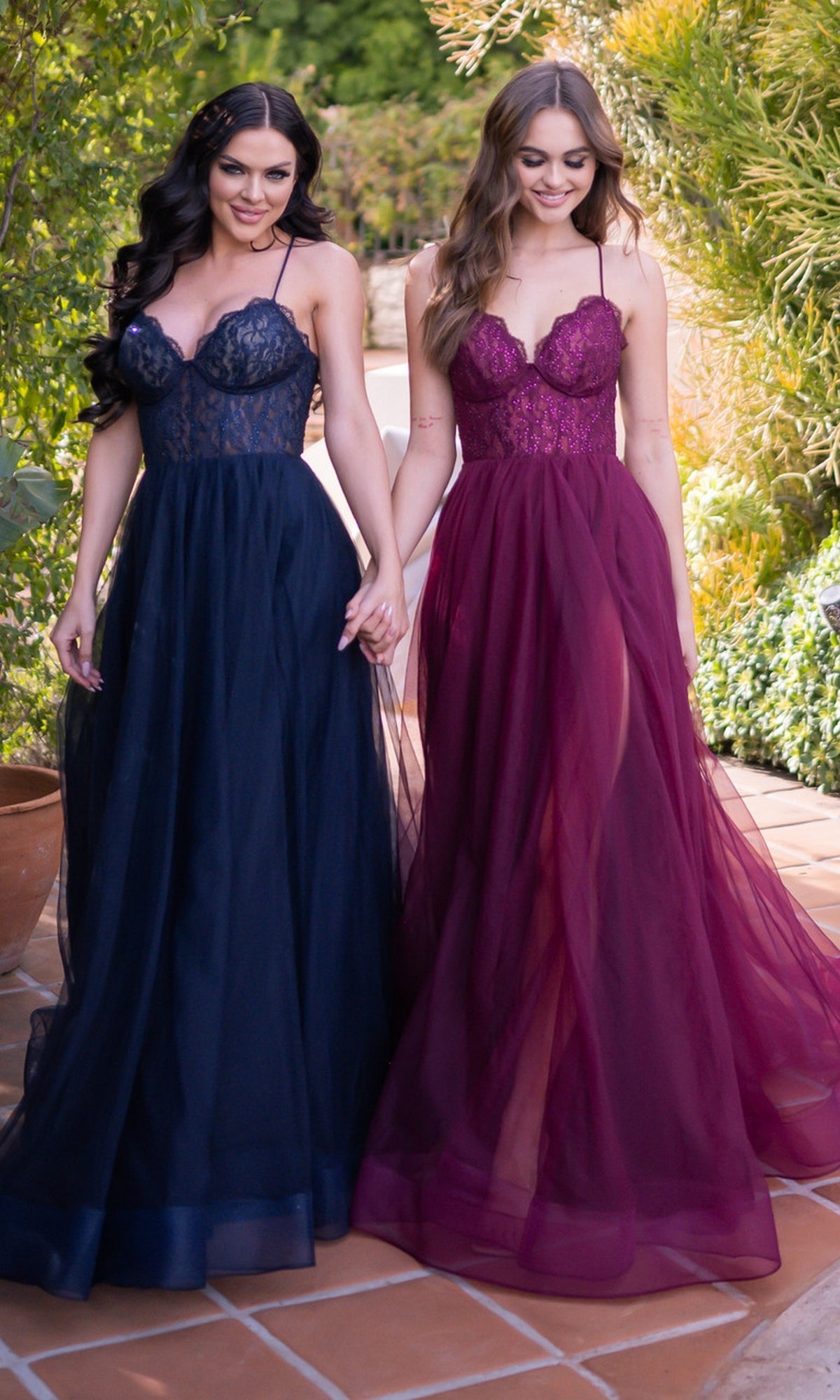 Two women stand outdoors holding hands, each wearing the Velvi Long Sweetheart A-Line Prom Dress Dani—one in navy blue and one in burgundy tulle—on a tiled pathway surrounded by greenery.