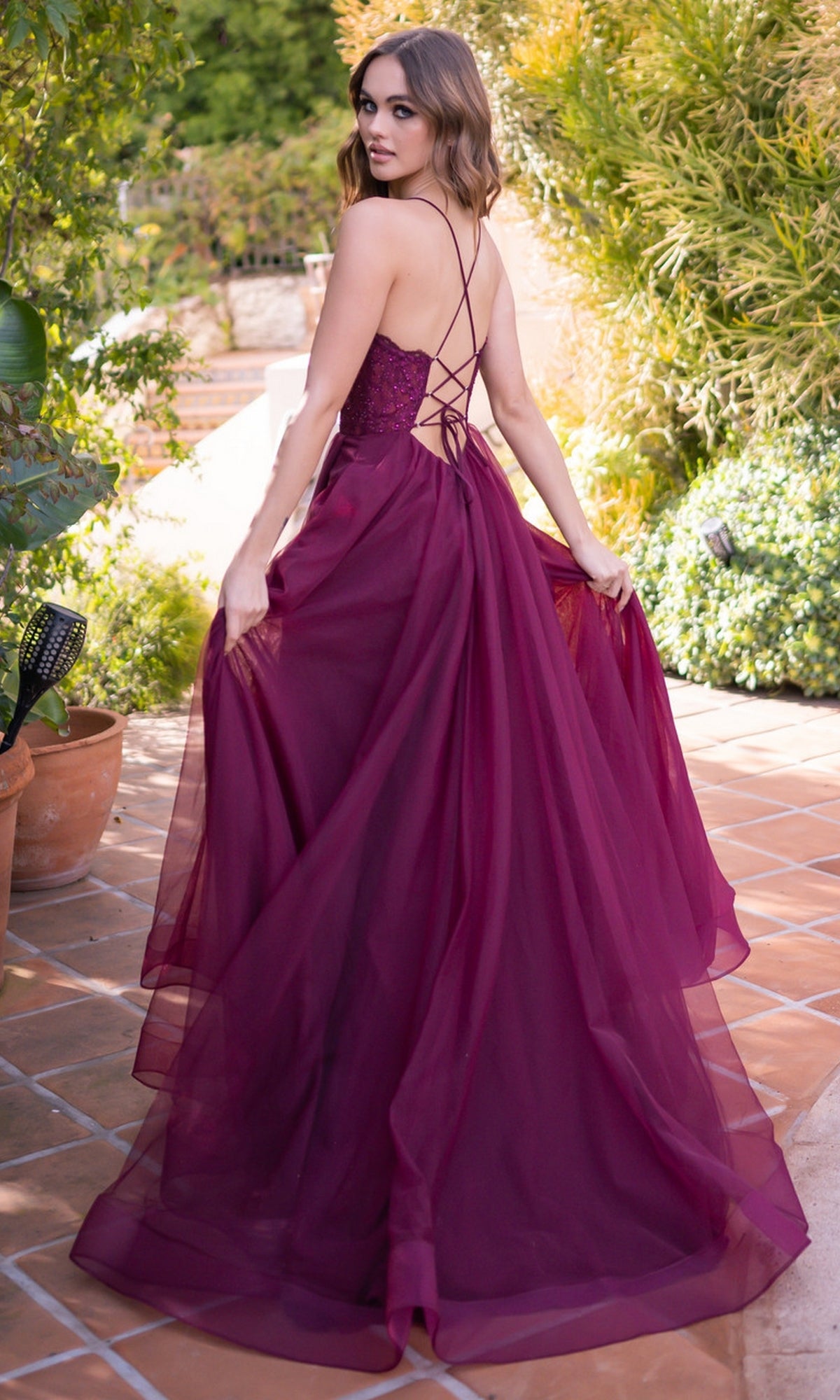 A woman in the Velvi Long Sweetheart A-Line Prom Dress Dani, a deep magenta, floor-length lace gown with a lace-up back, stands on a tiled patio holding her dress and looking over her shoulder.