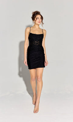 A woman stands against a plain background wearing the Short Homecoming Dress: Eureka EK122 in black, featuring thin straps, floral embroidery, an open back, and clear high-heeled shoes.