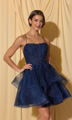 A woman wears the Short Homecoming Dress: Eureka EK137 in navy, featuring lace appliques and tulle, standing before a draped light yellow backdrop.