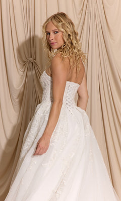 Woman with long curly blonde hair models the Long Wedding Dress: Eureka EK155 with detachable puff sleeves, standing turned in front of beige draped fabric.