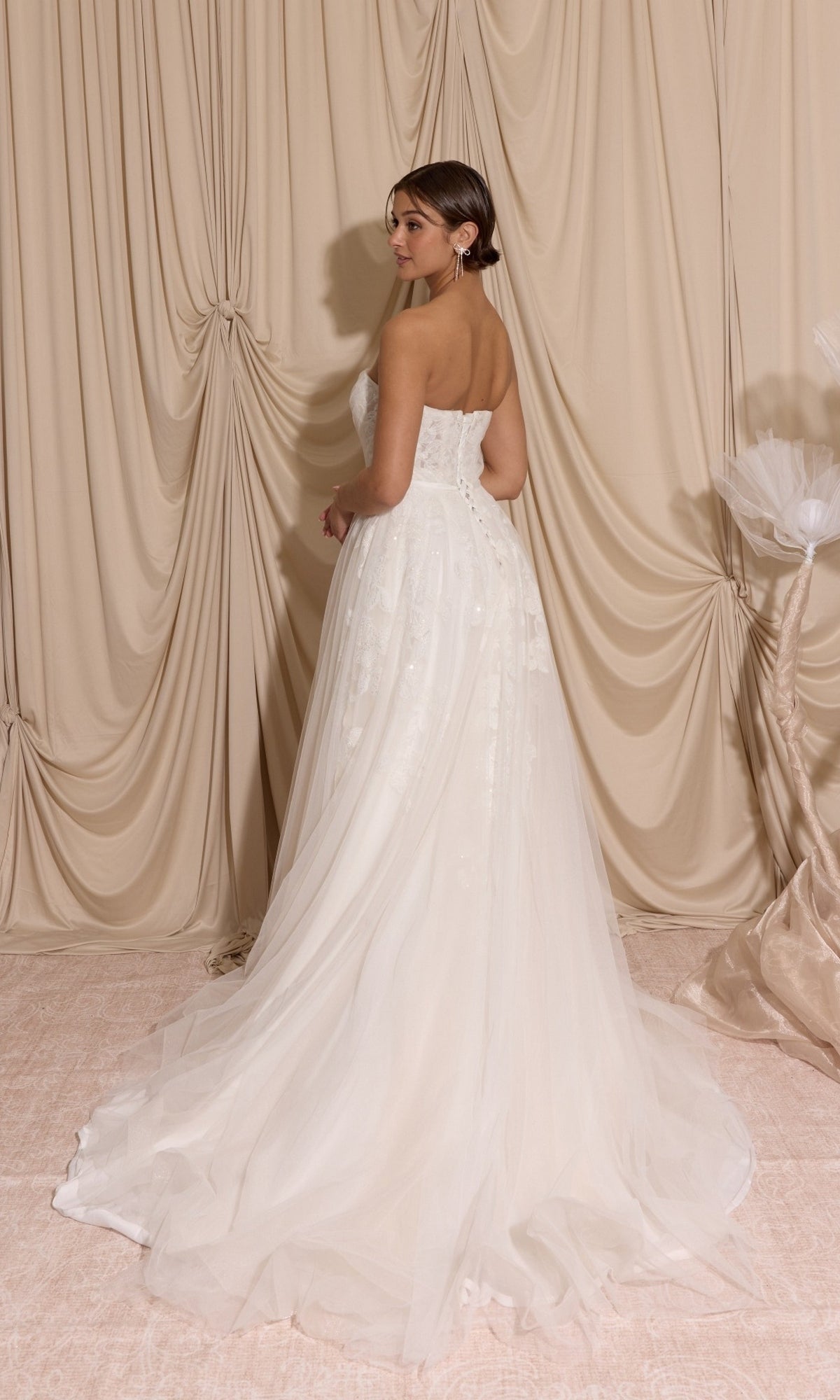 A woman wearing the Long Wedding Dress: Eureka EK165 with lace appliques and a tulle train stands before beige draped curtains, looking over her shoulder.