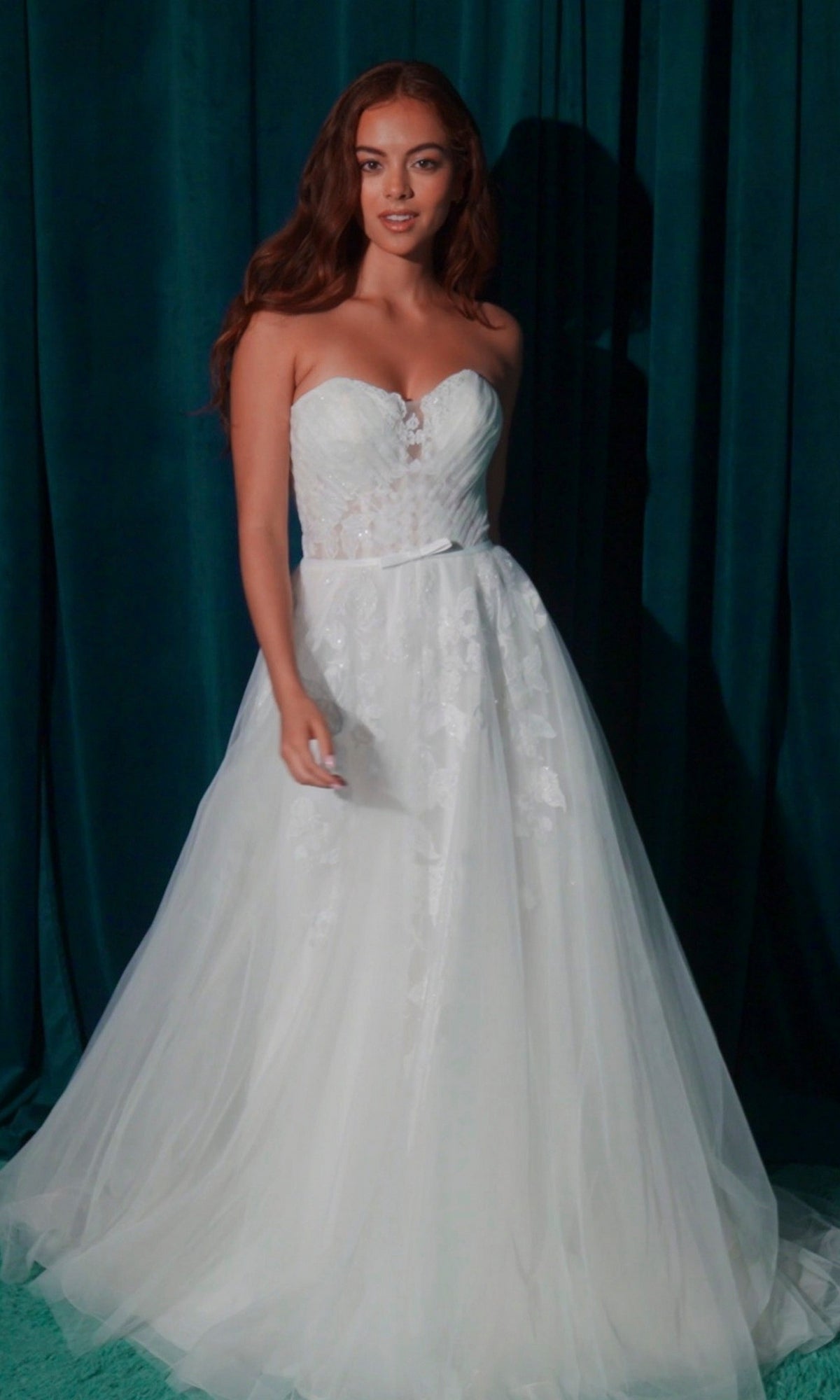 A woman stands before dark green curtains wearing the Long Wedding Dress: Eureka EK165, a strapless gown with lace appliques and a full tulle skirt.