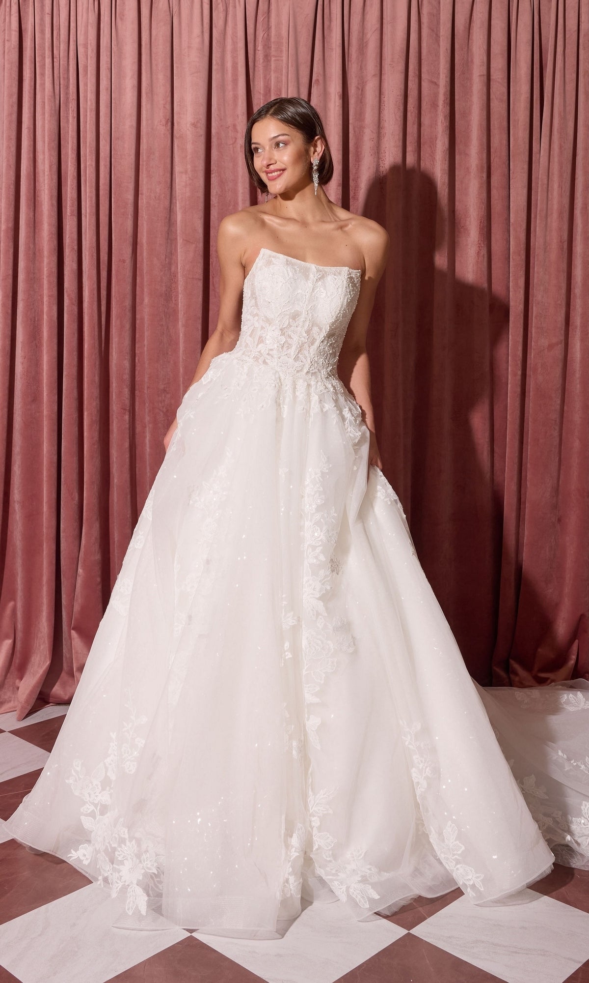 A woman in the Long Wedding Dress: Eureka EK175 with a strapless ball-gown silhouette stands on a checkered floor by pink curtains, looking aside and holding her dress.