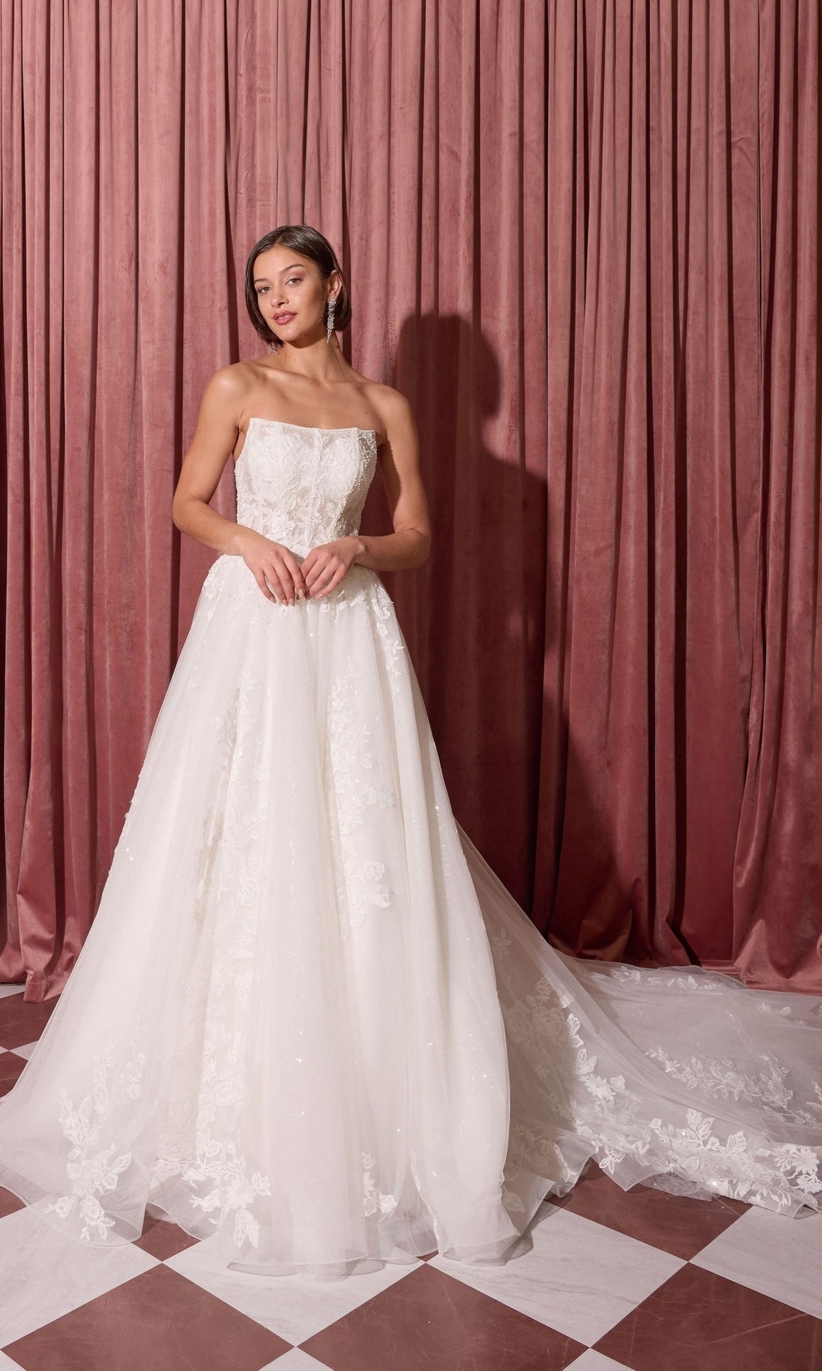 A woman in the Long Wedding Dress: Eureka EK175, off white, with a ball-gown skirt stands on a checkered floor before pink curtains.