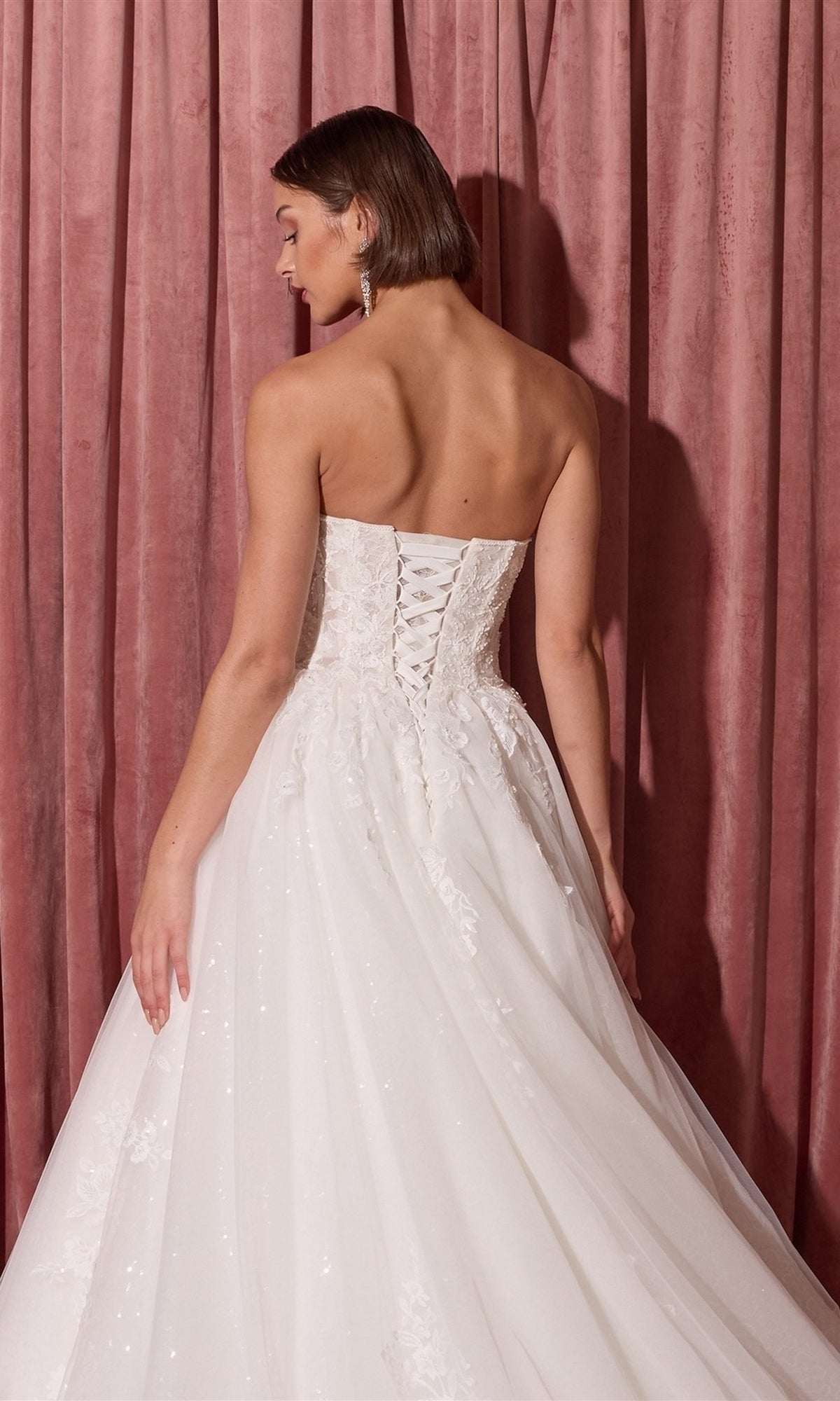 A woman wearing the Long Wedding Dress: Eureka EK175 with a lace-up back and ball-gown skirt stands before pink curtains, facing away from the camera.