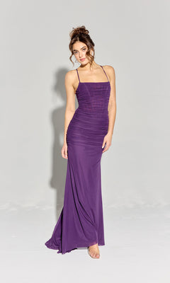 A woman stands in a studio wearing the Eureka EK177 Strappy-Back Slinky Long Prom Dress, a sleeveless purple gown with thin straps and ruched detailing, paired with open-toe heels.