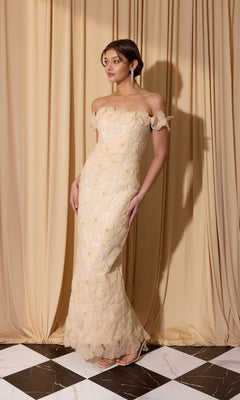 A woman stands on a black and white checkered floor in front of beige curtains, wearing the Eureka EK205 Long Prom Dress in cream, its elegant off-the-shoulder lace design flowing gracefully to the floor.