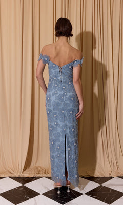 A person stands on a black and white tile floor, facing beige curtains, wearing the Eureka EK205 long prom dress in light blue floral lace with off-the-shoulder sleeves, a lace-up back, and a slit at the hem.