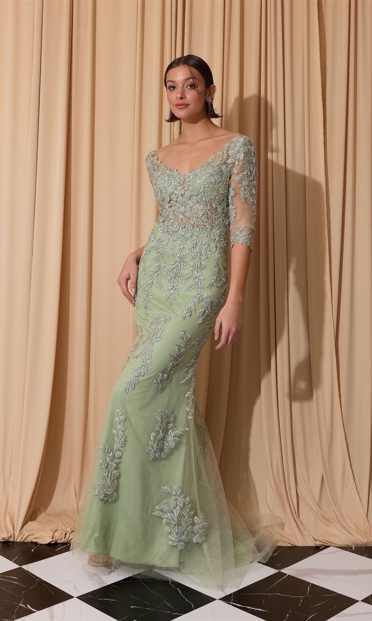 A woman stands on a black-and-white checkered floor in the Long Formal Dress: Eureka EK209, a fitted sage mermaid gown with sheer sleeves and silver floral embroidery, before beige curtains.