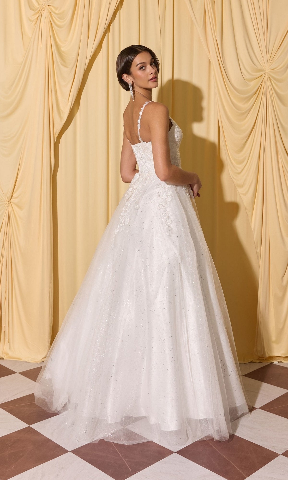 Woman in a white Long Prom Dress: Eureka EK210 with 3D floral appliques stands on a checkered floor, facing away, with yellow curtains behind her.