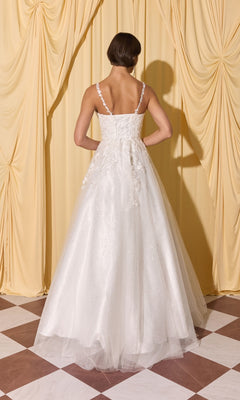 A woman faces yellow curtains with her back to the camera, wearing the Long Prom Dress: Eureka EK210, a white floor-length gown with 3D floral appliques.