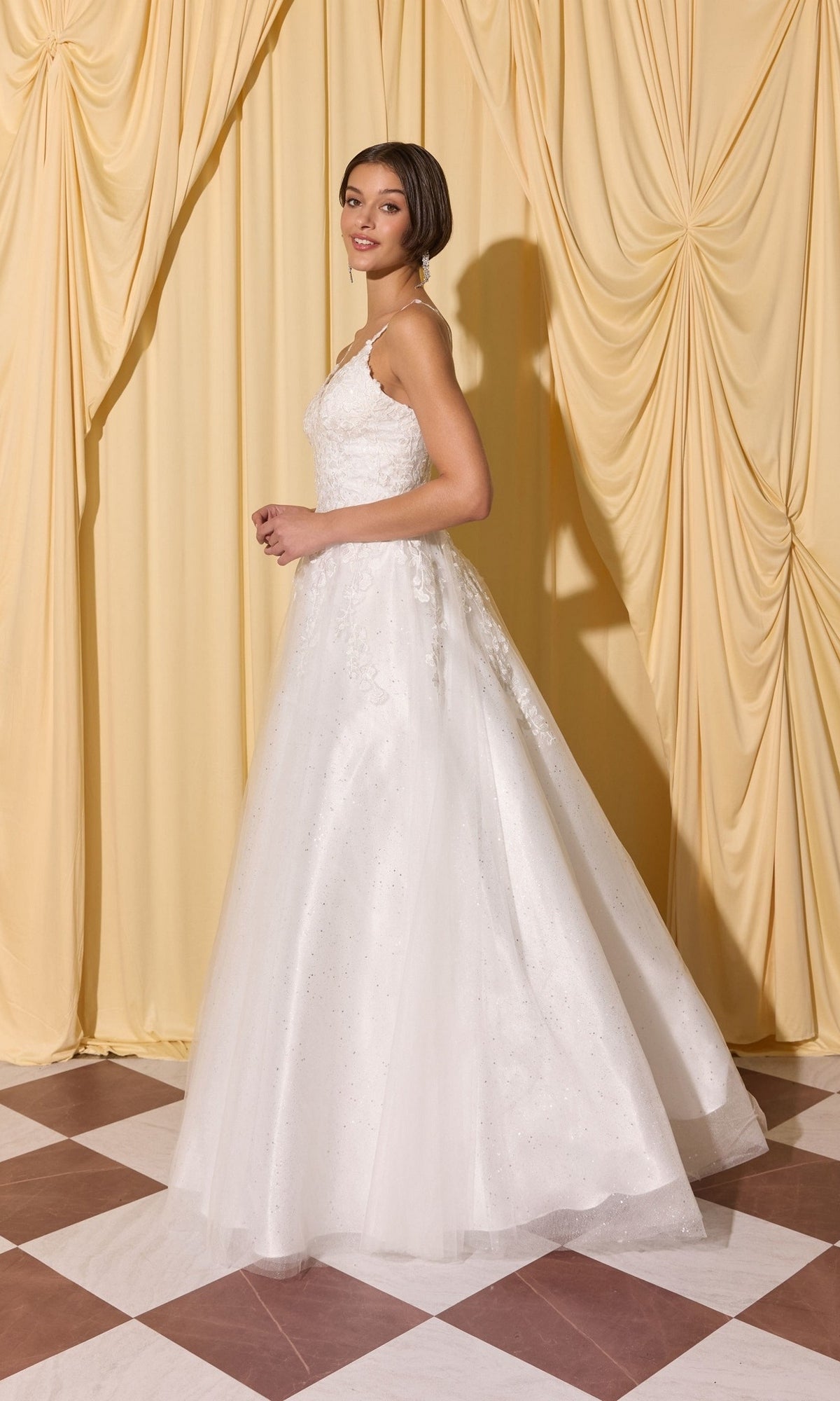 A woman in the Long Prom Dress: Eureka EK210, a white sleeveless gown with 3D floral appliques, stands on a checkered floor before yellow drapes, looking over her shoulder.