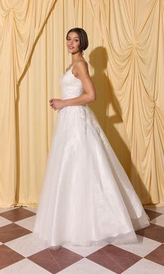 A woman in the Long Prom Dress: Eureka EK210, a white sleeveless gown with 3D floral appliques, stands on a checkered floor before yellow drapes, looking over her shoulder.