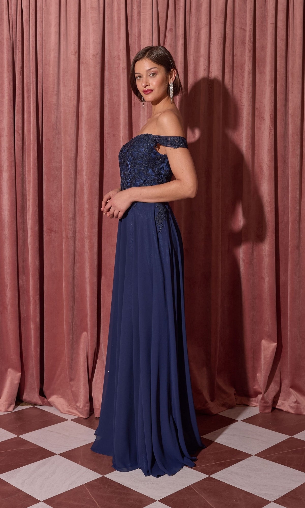 Woman in the Long Prom Dress: Eureka EK211 with a lace applique bodice stands on a checkered floor before pink curtains, posing with hands clasped.