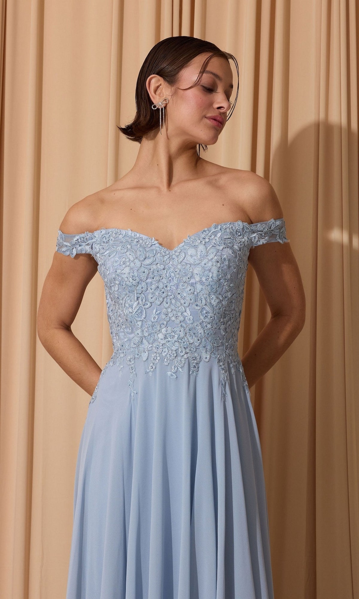 A woman in the Long Prom Dress: Eureka EK211, featuring an off-the-shoulder lace applique bodice and pockets, stands against beige curtains.