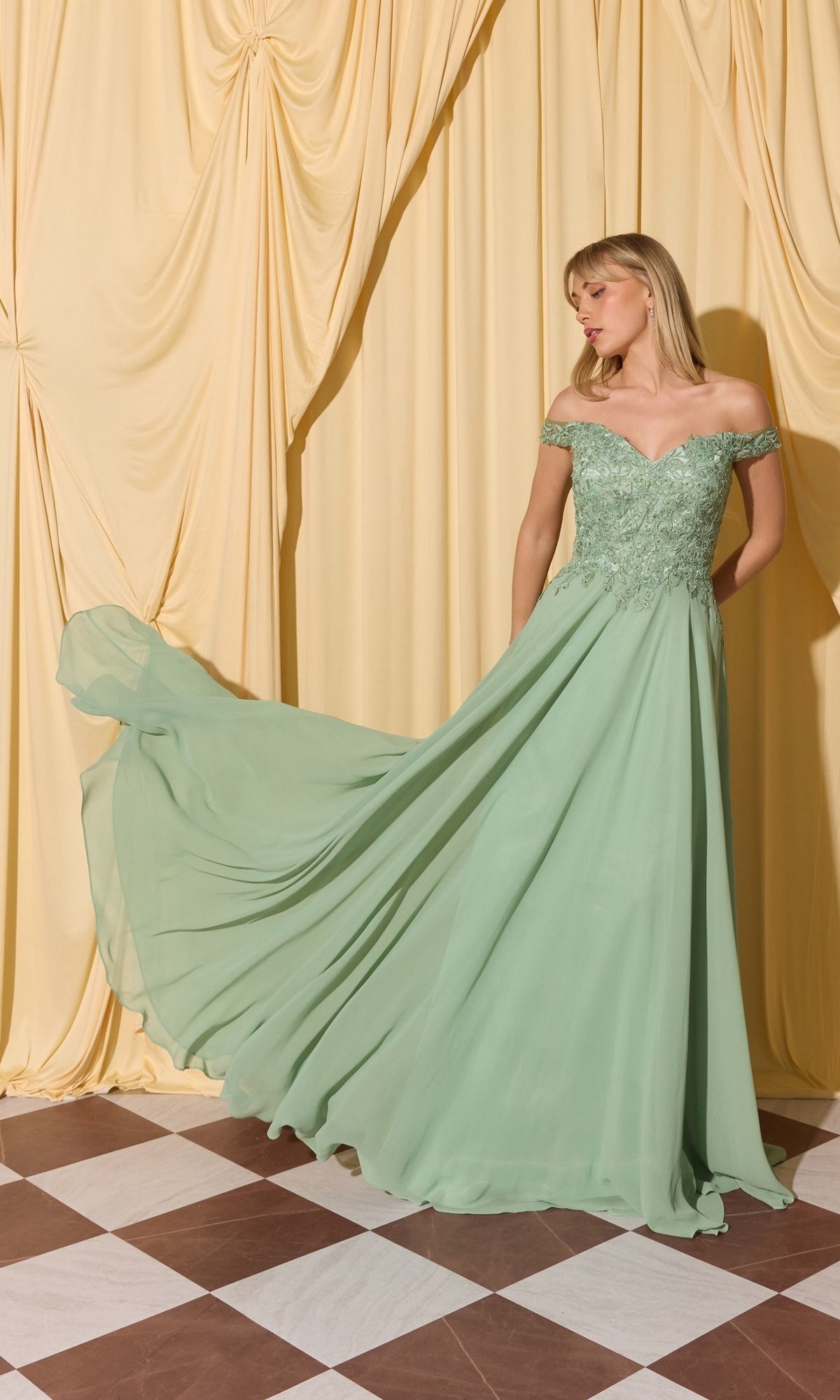 A woman in the Long Prom Dress: Eureka EK211, a light green off-the-shoulder gown with lace applique bodice, stands before cream drapes on a checkered floor.