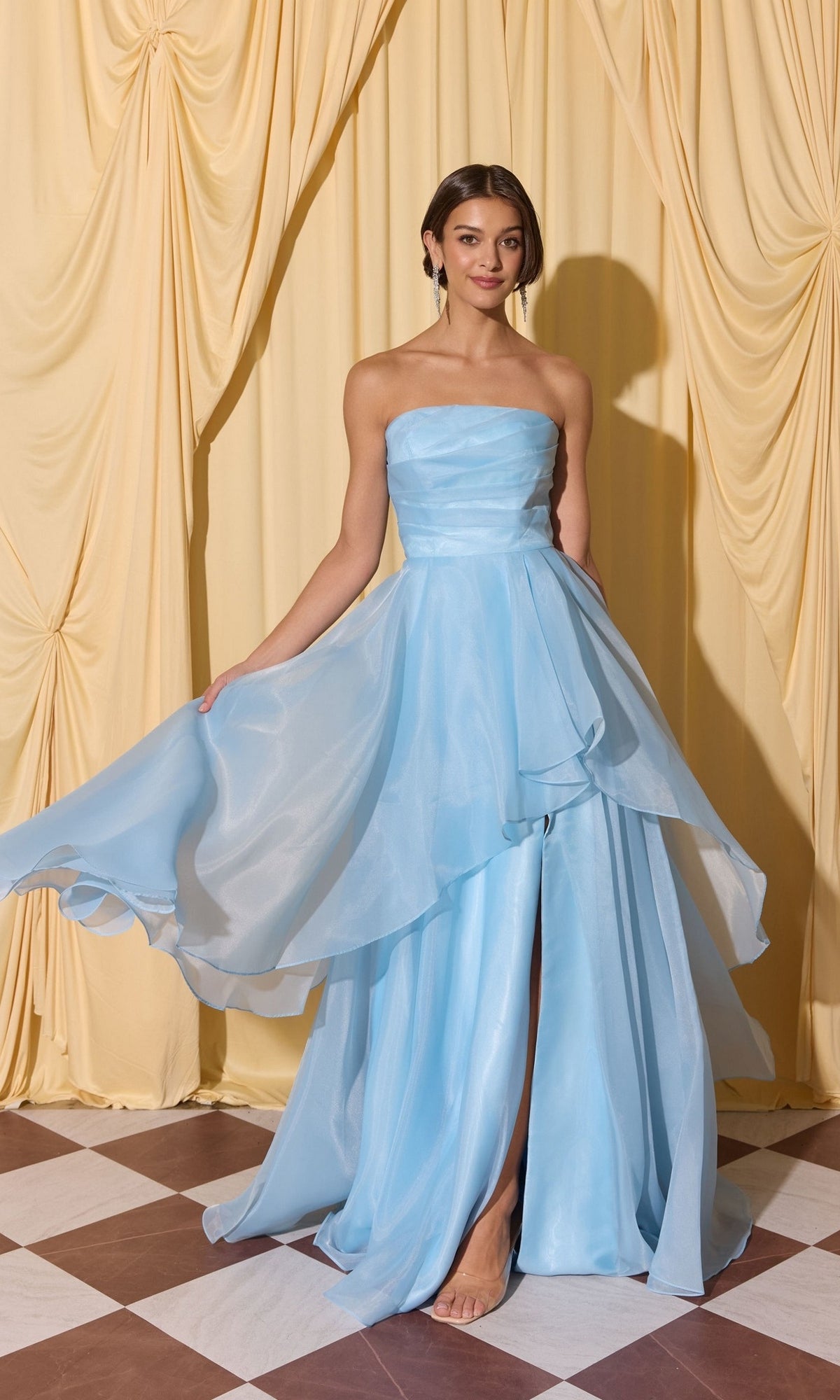 A woman wearing the Long Prom Dress: Eureka EK216 with an a-line skirt stands barefoot on a checkered floor, holding her layered gown before cream-colored curtains.