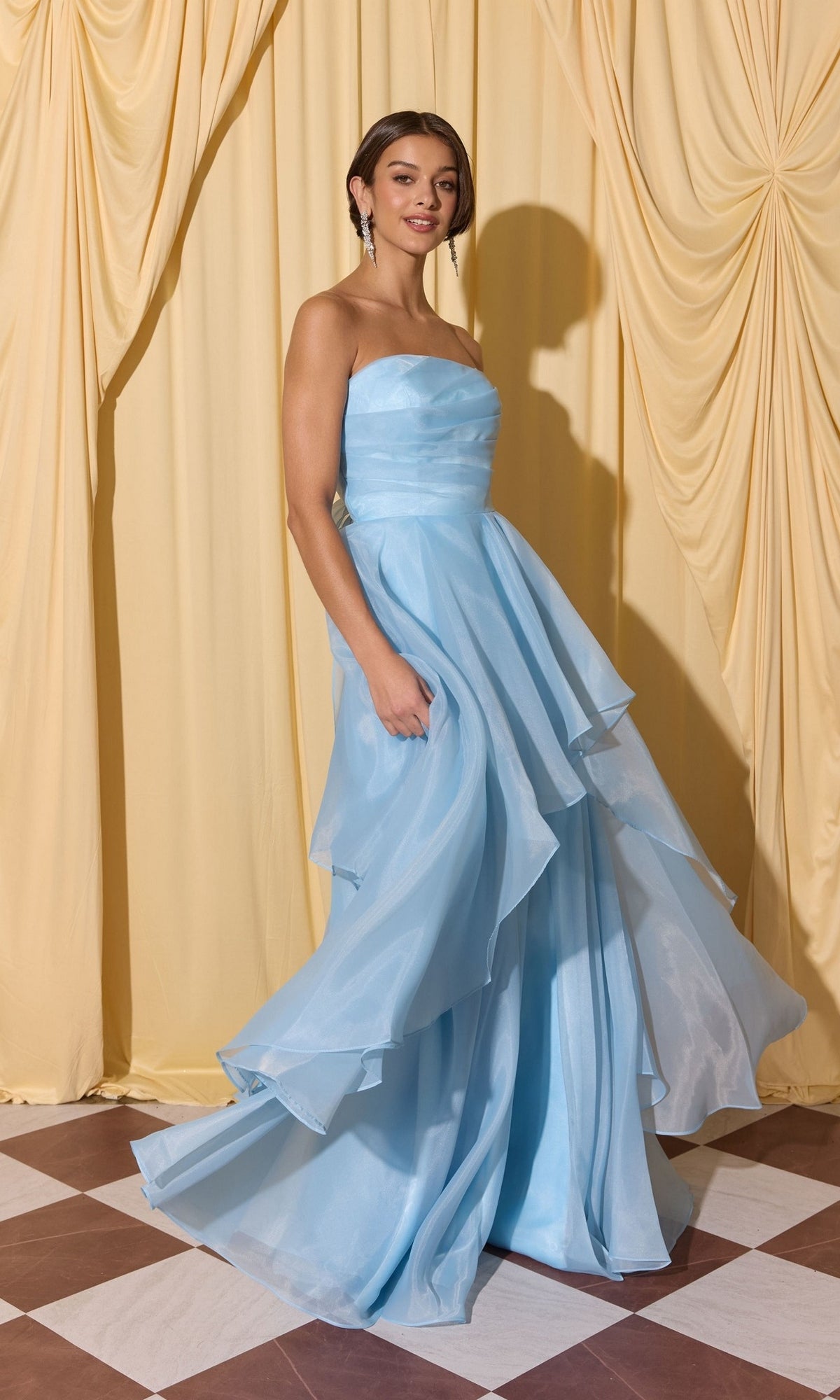 A woman wearing the Long Prom Dress: Eureka EK216, a strapless gown with an a-line skirt, stands on a checkered floor before yellow draped curtains.