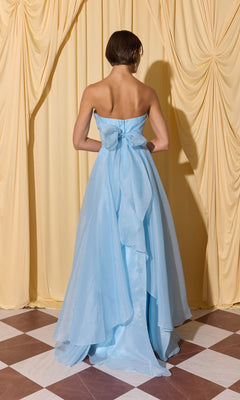 A woman wearing the Long Prom Dress: Eureka EK216, featuring a strapless bodice, large bow, and flowing a-line skirt, stands on a checkered floor before yellow curtains.