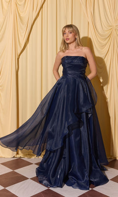 A woman in the Long Prom Dress: Eureka EK216, navy color, with an a-line skirt, stands on a checkered floor before yellow drapes, holding one side of her dress.