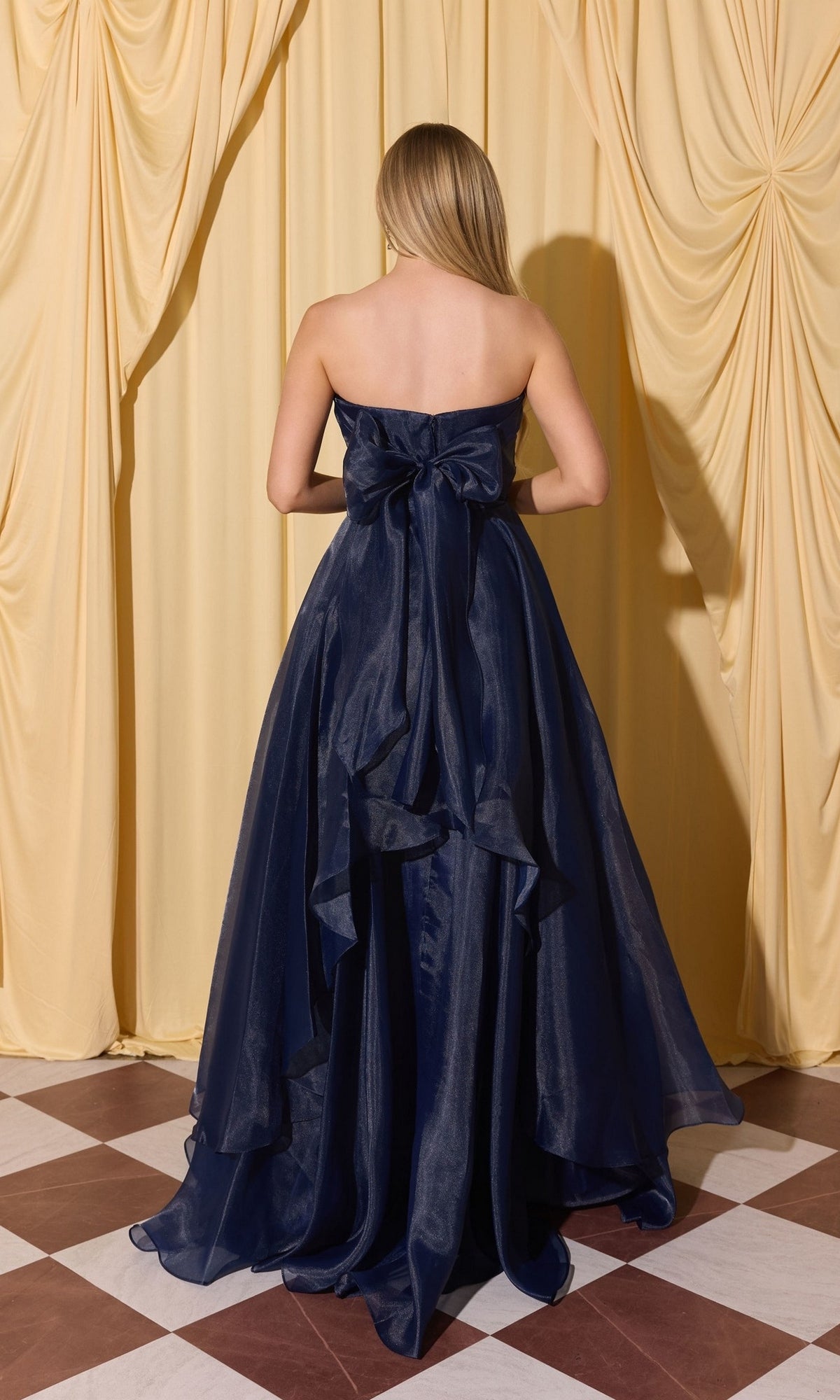 A woman in the Long Prom Dress: Eureka EK216, featuring a strapless design and large back bow, stands before yellow draped curtains on a checkered floor.