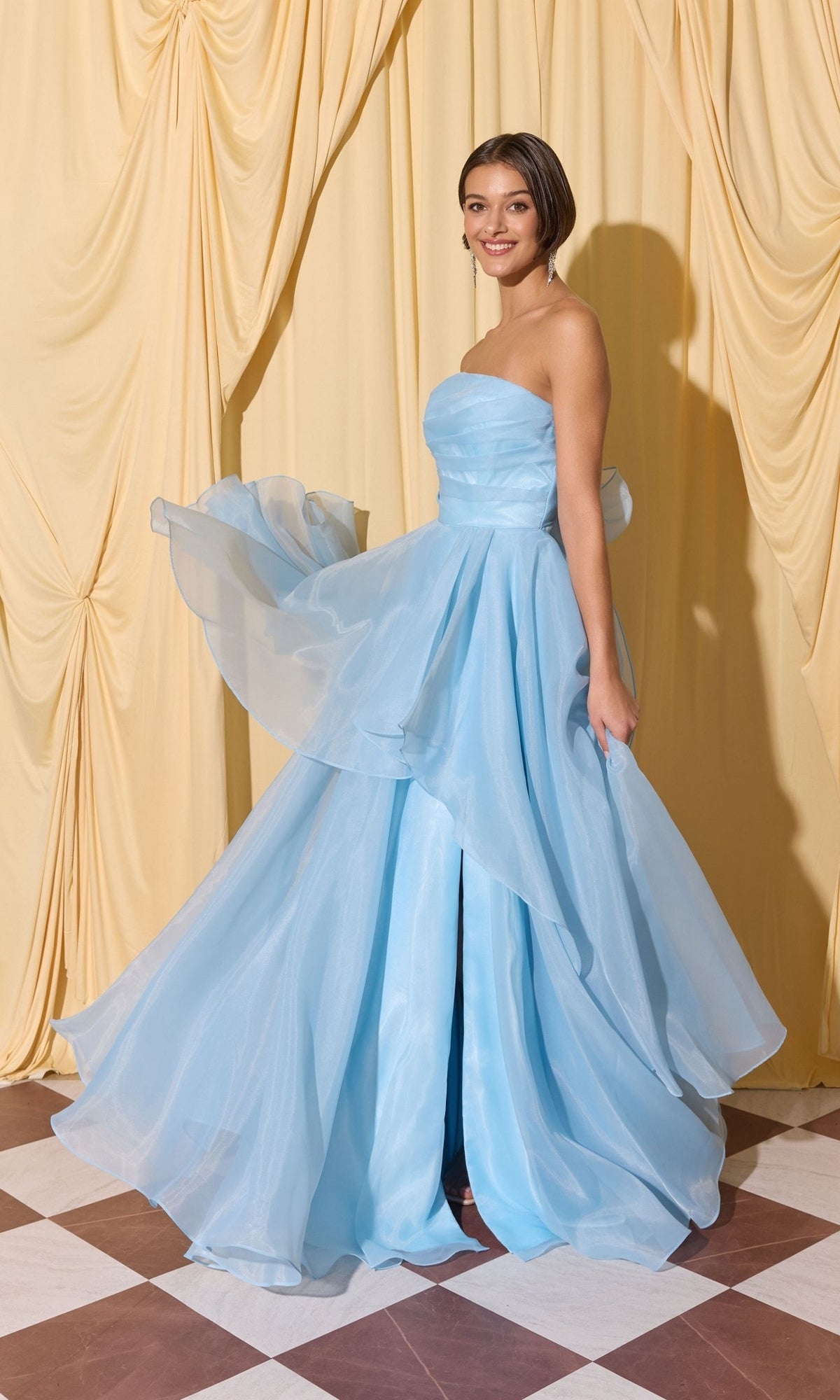 Smiling at the camera, a woman in the Long Prom Dress: Eureka EK216 in powder blue stands on a checkered floor before gathered cream curtains.