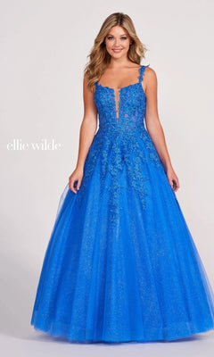 A woman in the Ellie Wilde EW120014 Long Glitter Prom Ball Gown, a bright blue floor-length A-line dress with floral lace appliqués and a glitter tulle skirt, stands against a plain light background. "ellie wilde" appears on the left.