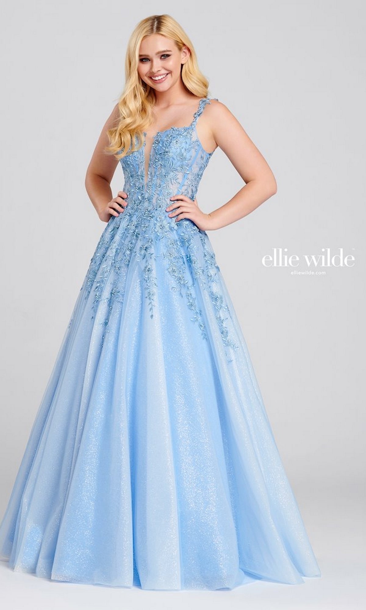 A woman smiles at the camera wearing the Ellie Wilde EW120014 long glitter prom ball gown in light blue, featuring floral embroidery, a deep V neckline, and floor-length tulle.