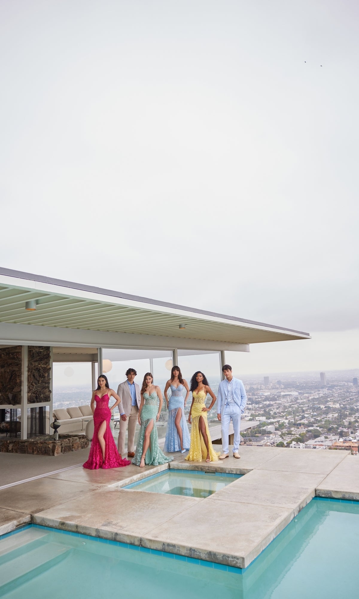 Six people in formalwear, including one wearing the Ellie Wilde Long Lace Mermaid Prom Dress EW35005, stand by a rooftop pool with a cityscape behind them.