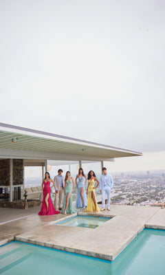 Six people in formalwear, including one wearing the Ellie Wilde Long Lace Mermaid Prom Dress EW35005, stand by a rooftop pool with a cityscape behind them.