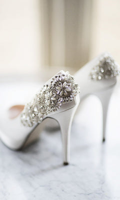 The Indulgence Ivory Satin Prom Shoes: Paradox London P1710 feature jeweled heels and ornate silver floral embellishments, elegantly displayed on a marble surface.