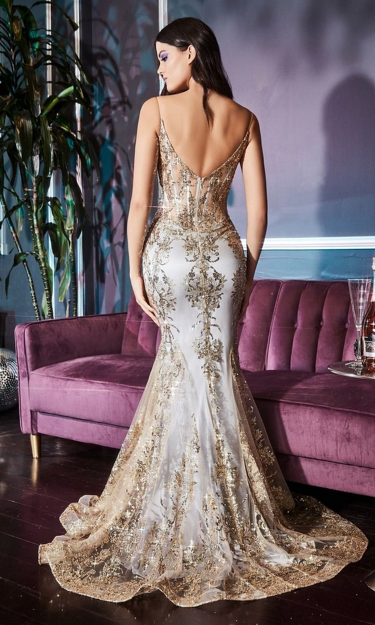 Wearing the Glitter-Print Long V-Neck Mermaid Prom Dress J810A, a woman with a corset-style bodice and intricate gold and white glitter details stands before a purple sofa in a stylish room.