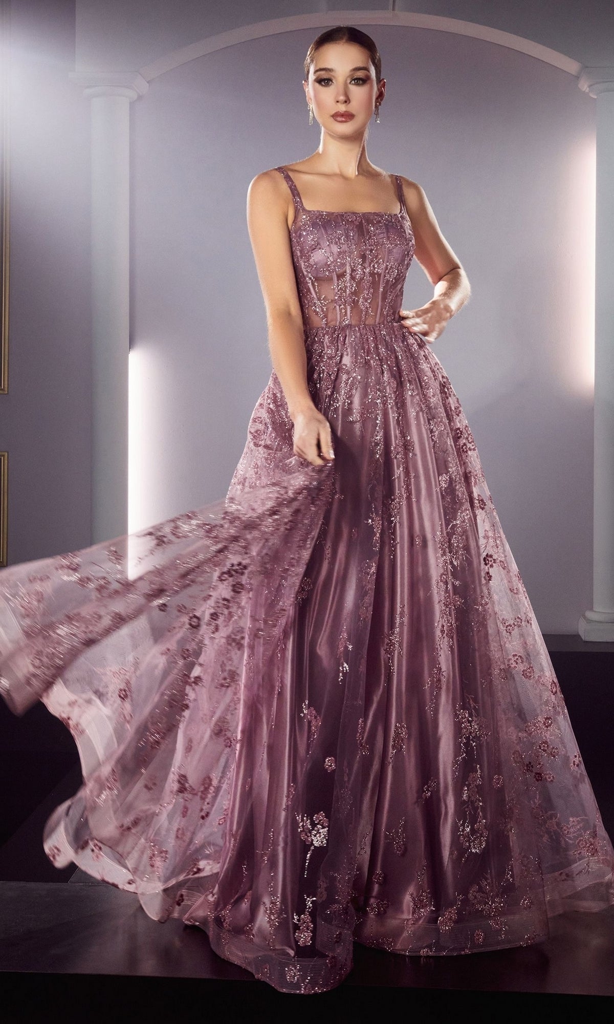 A woman poses in the Long A-Line Glitter-Print Prom Dress J840, a floor-length gown featuring sheer panels and intricate floral detailing, set against a backdrop with arched columns.