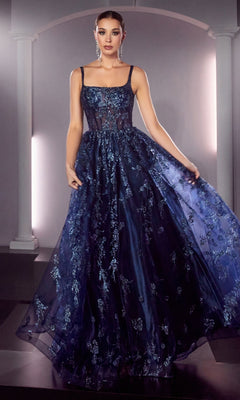 A woman stands in the Long A-Line Glitter-Print Prom Dress J840, featuring a sleeveless navy blue design with floral embroidery and a sheer bodice, holding her skirt before an arched architectural backdrop.