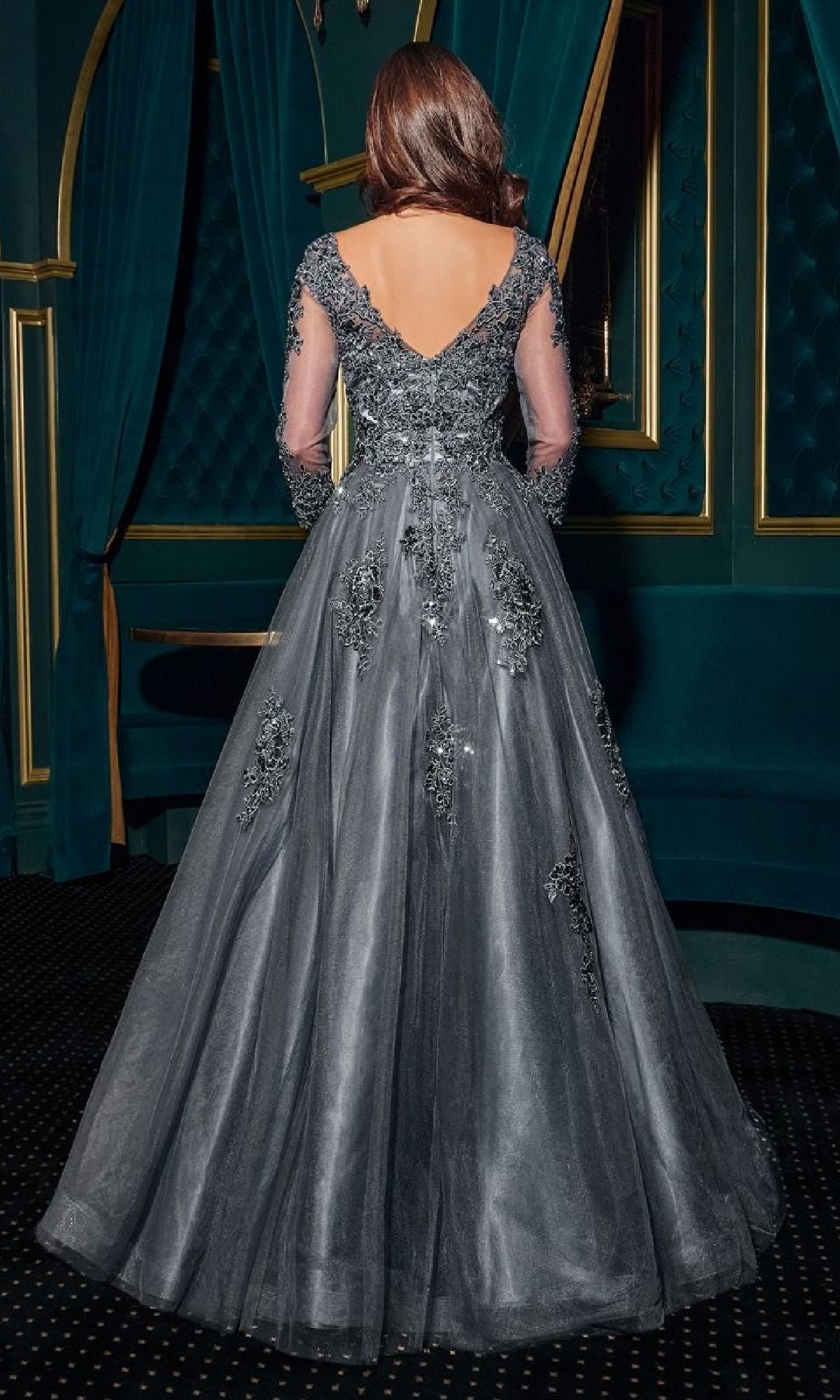 A woman with long brown hair wears the Ladivine J883, a long sleeve formal dark gray gown with beaded details and a deep V-back, standing in an ornate room with teal walls and drapes.