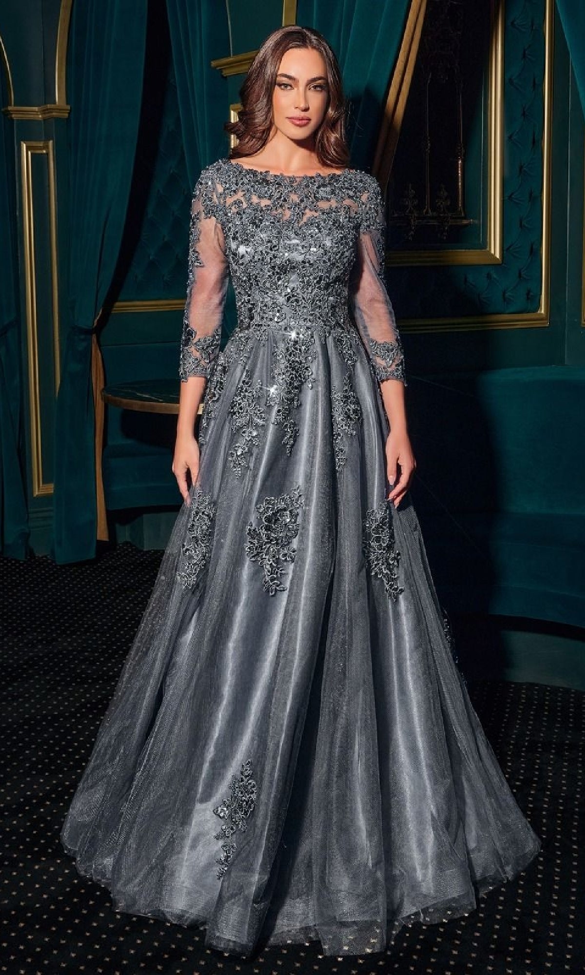 A woman in the Ladivine J883 long sleeve formal dress with silver-gray lace and sequin details stands in a formal room with dark teal curtains and gold trim.