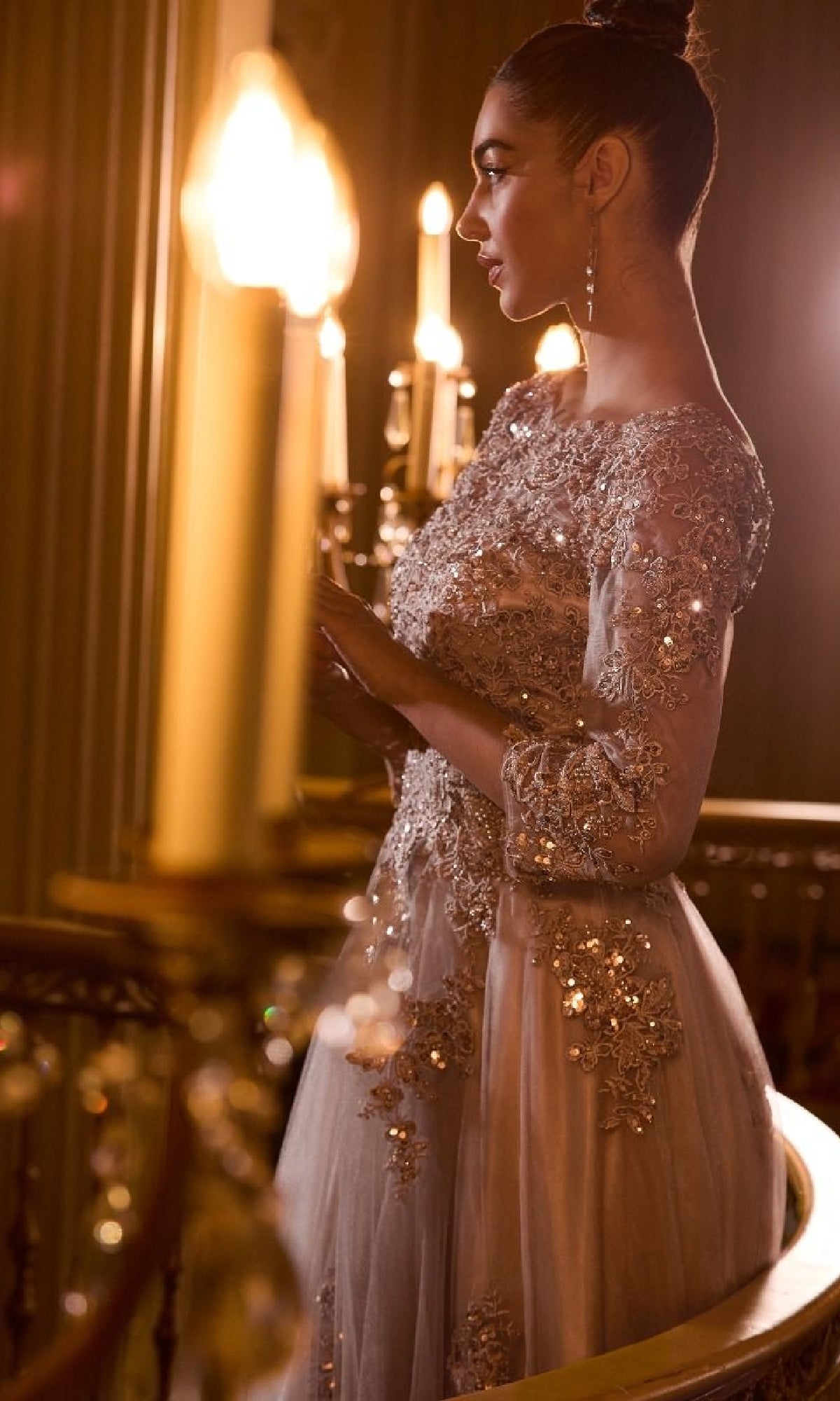 A woman in a Long Sleeve Long Formal Dress: Ladivine J883 stands by a railing under warm chandelier light, her hair styled in a bun and accessorized with long earrings.