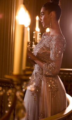 A woman in a Long Sleeve Long Formal Dress: Ladivine J883 stands by a railing under warm chandelier light, her hair styled in a bun and accessorized with long earrings.