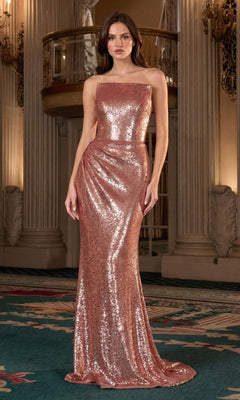 A woman stands indoors wearing the Ladivine J904, a floor-length rose gold prom dress with sequins and a square neckline. Ornate columns and railings can be seen in the background.