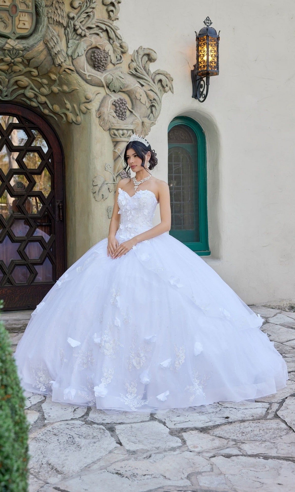 A woman wearing the Long Wedding Dress: Juliet JT1495HW in white stands on a stone path before an ornate doorway with a curved, vine-like design and a green-framed window.