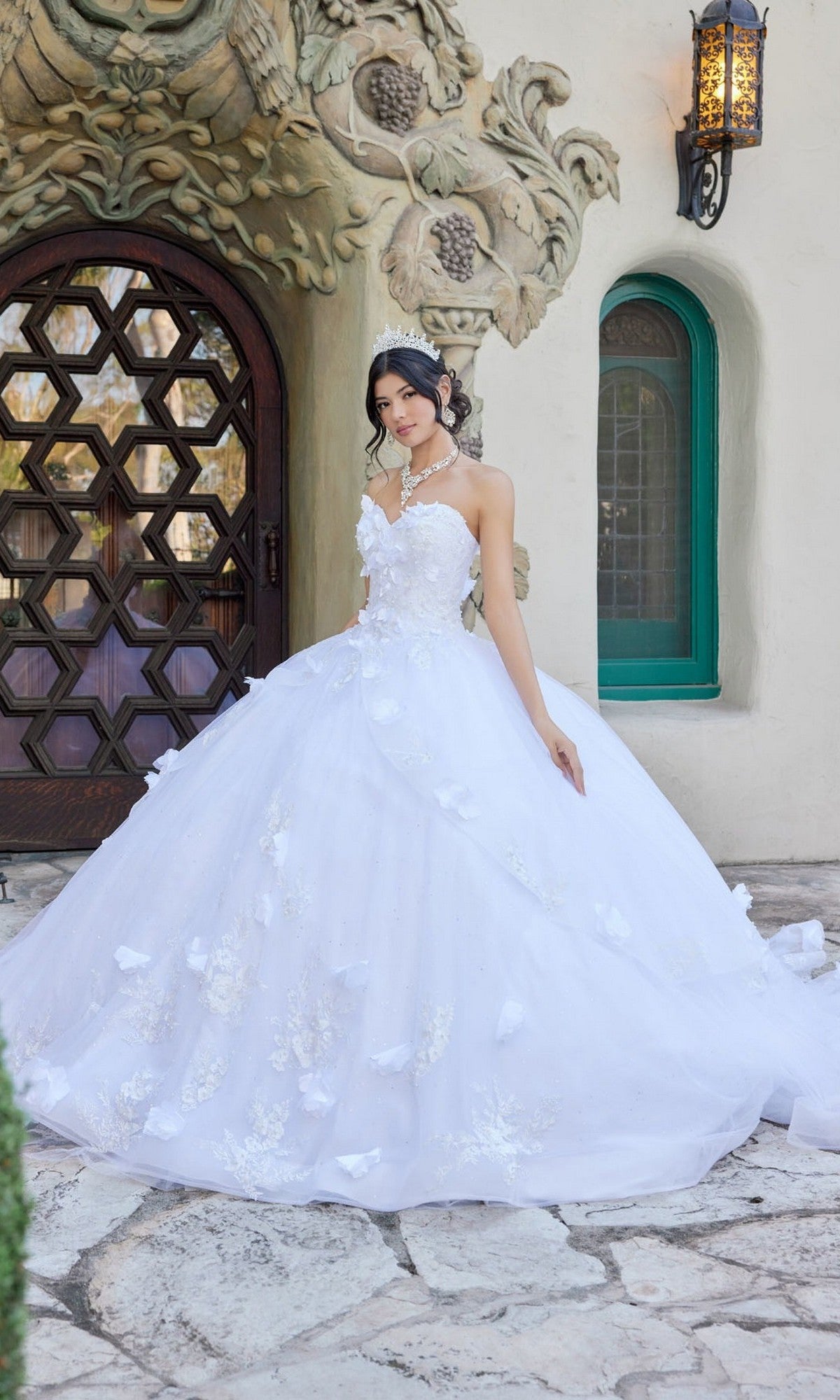 Wearing the Long Wedding Dress: Juliet JT1495HW—a strapless white ball gown featuring a sweetheart neckline and delicate floral appliques—a woman stands outside an ornate building with carved details, posing for a formal portrait.