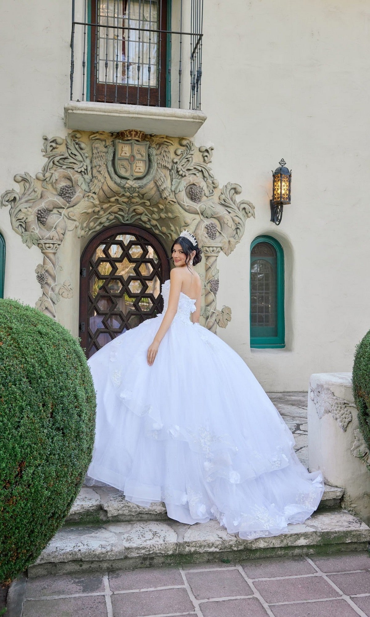 A young woman in the Long Wedding Dress: Juliet JT1495HW, a strapless white ball gown with a sweetheart neckline, stands on stone steps in front of an ornate arched doorway, looking over her shoulder.