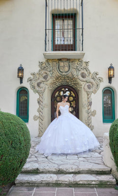 Wearing the Juliet JT1495HW long wedding dress, which features a strapless white ball gown design with delicate floral appliques, a person stands before an ornate doorway framed by lush greenery and rounded shrubs.