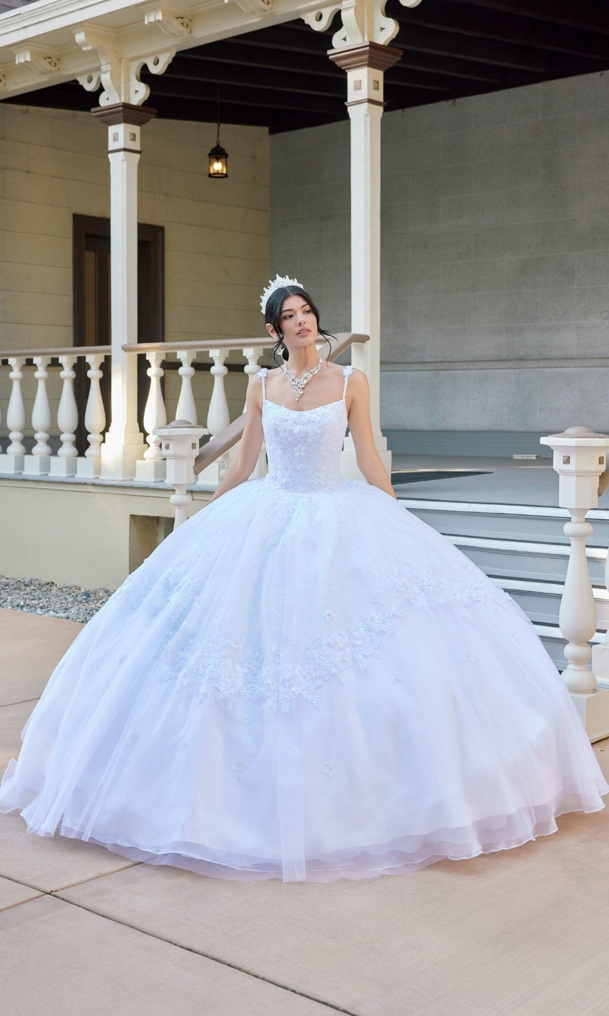 A woman wearing the Long Wedding Dress: Juliet JT1496HW in white, adorned with delicate floral lace appliques and a tiara, poses outdoors in front of a porch with white railings and steps.