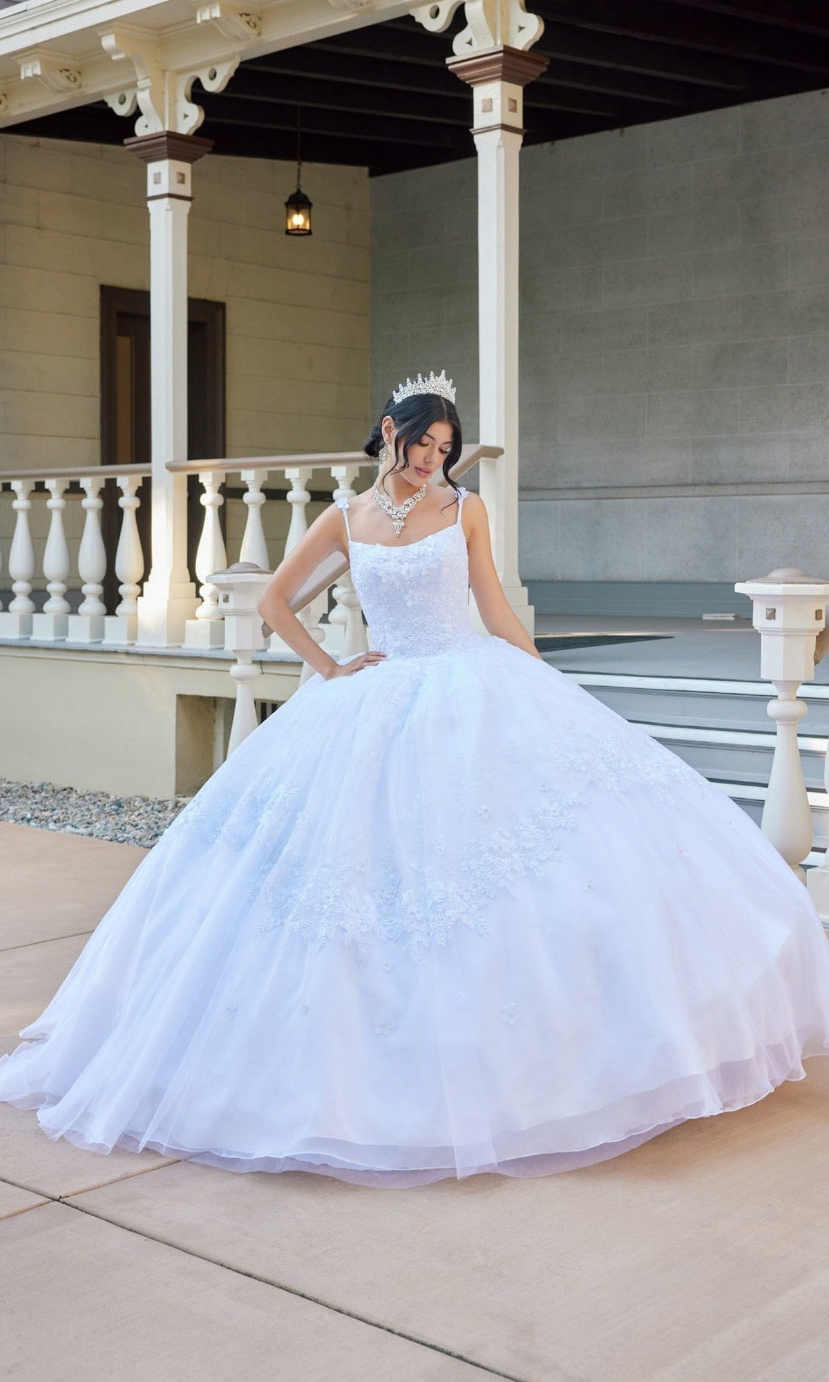 A woman wearing the Long Wedding Dress: Juliet JT1496HW, a white gown adorned with delicate floral lace appliques, and a shimmering tiara stands on a paved area in front of a porch with white railings and columns, looking down at her elegant dress.