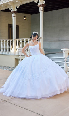 A woman wearing the Long Wedding Dress: Juliet JT1496HW, a white gown adorned with delicate floral lace appliques, and a shimmering tiara stands on a paved area in front of a porch with white railings and columns, looking down at her elegant dress.