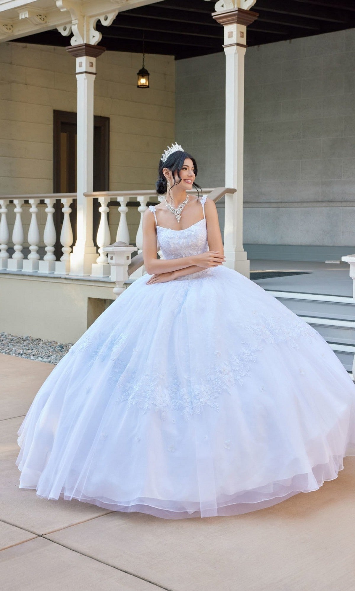 Wearing the Long Wedding Dress: Juliet JT1496HW, a young woman in a tulle ball gown adorned with delicate floral lace appliques and a sparkling tiara stands on a porch, gazing to the side with her hands clasped.
