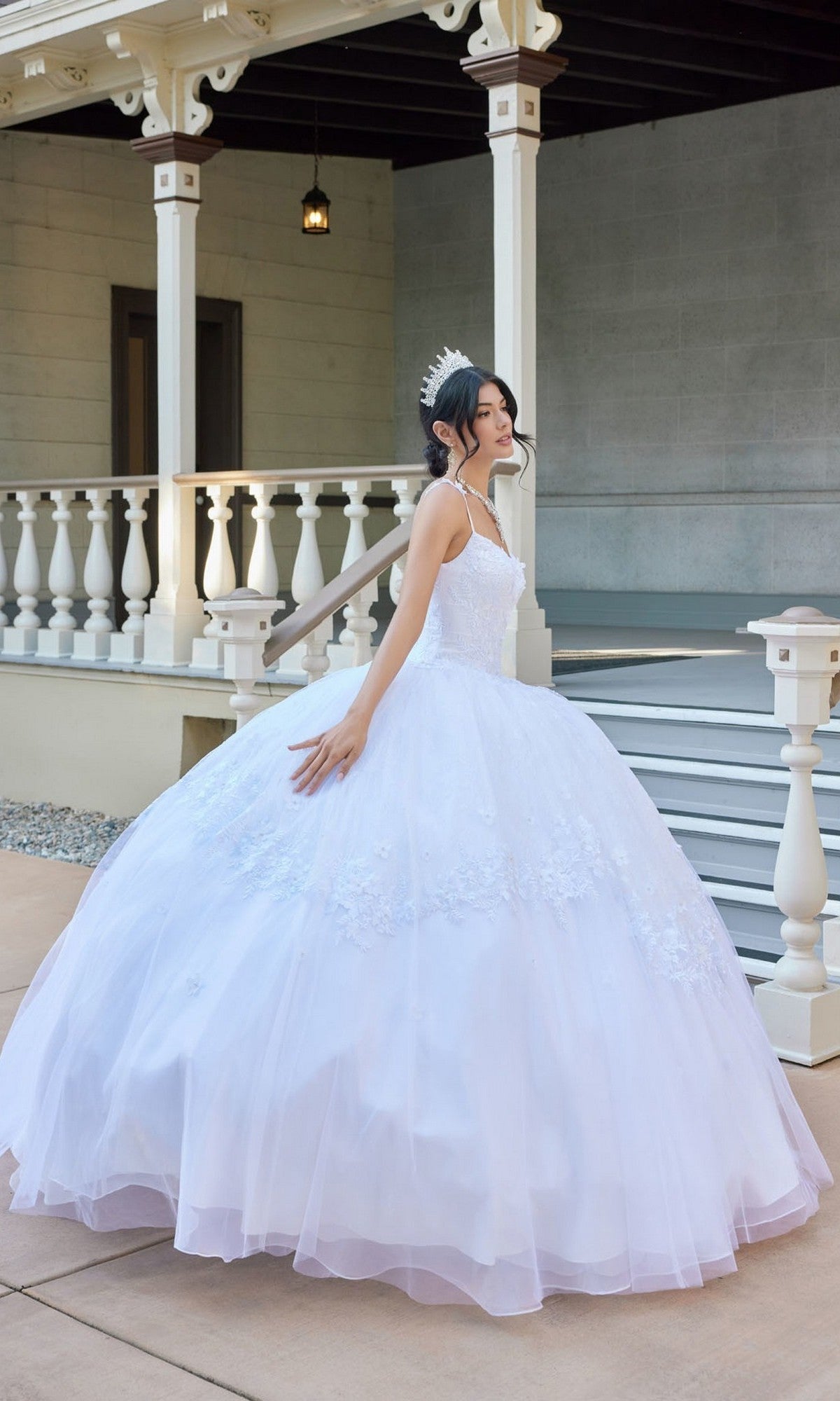 A young woman stands to the side in front of a porch with white railings and steps, wearing the Juliet JT1496HW Long Wedding Dress—a white tulle ball gown adorned with delicate floral lace appliques—paired with a sparkling tiara.